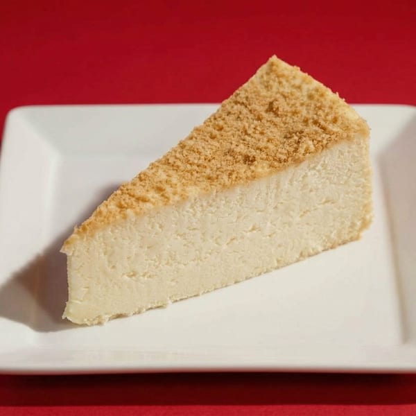A slice of cheesecake with a crumbly topping sits on a white rectangular plate against a red background.