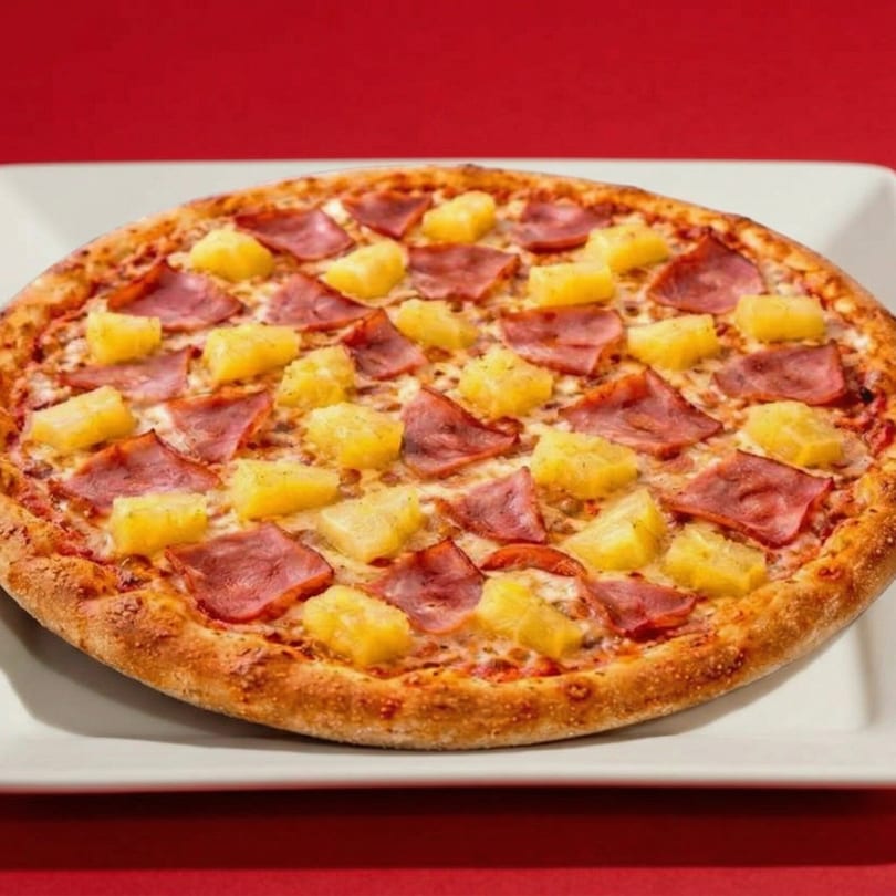 A whole Hawaiian pizza with ham and pineapple on a white plate against a red background.